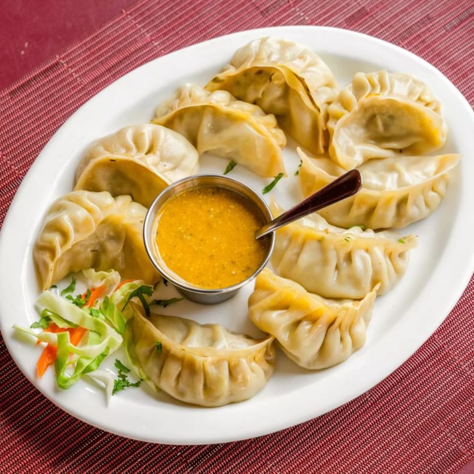 A white plate of Himalayan momos, a small bowl of orange dipping sauce, and a side salad.