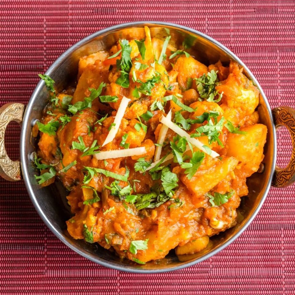 A close-up of Indian curry with potatoes and cilantro in a metal bowl on a red mat.