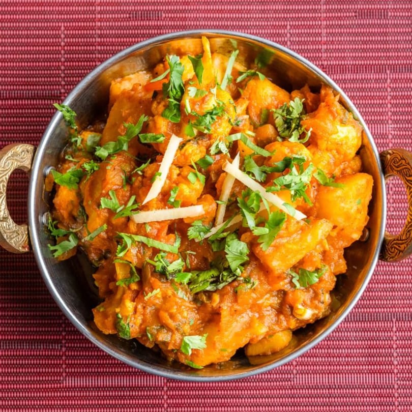 A close-up of Indian curry with potatoes and cilantro in a metal bowl on a red mat.